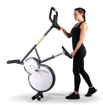 A fitness instructor holding a stylish modern exercise bike with a sleek design, featuring three adjustable grip positions and a multifunctional console. Stil-Fit Ergometer Pure - Stationary Bike